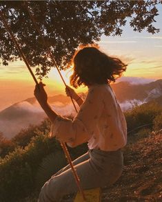 a woman is sitting on a swing in the mountains at sunset with her hair blowing in the wind