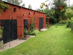 a red building with black doors in the middle of a grassy area next to trees and bushes