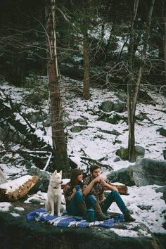 a man and woman are sitting on a blanket in the snow next to a white dog