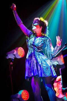 a woman is performing on stage with her arms in the air and lights behind her