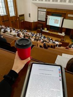 a person holding a coffee cup and tablet in front of an auditorium full of people