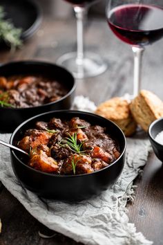 two black bowls filled with stew next to wine glasses and bread on a wooden table