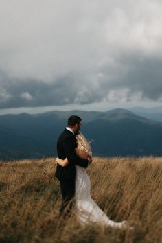 a bride and groom standing in tall grass on top of a hill with mountains in the background