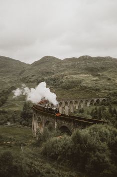 a train traveling over a bridge on top of a lush green hillside next to mountains