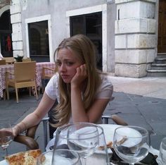 a woman sitting at an outdoor table with pizza and wine glasses on it, talking on the phone