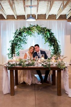 a bride and groom sitting at a table with greenery on it, surrounded by candles