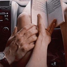 two hands holding an open book while sitting in the driver's seat of a car