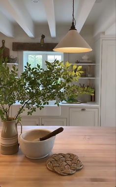 a bowl and spoon on a wooden table in a room with white cabinets, windows, and potted plants