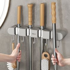 two hands holding brooms and brushes in front of a wall mounted rack with hooks