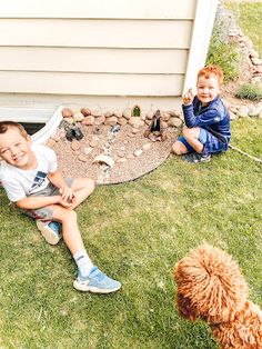 two young boys sitting on the ground next to a brown dog and a white house