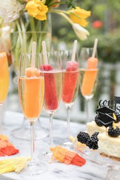 several glasses filled with different types of drinks on a table next to flowers and fruit