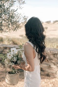 a woman holding a bouquet of flowers in front of an olive tree with her back to the camera