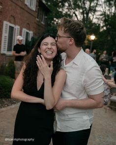 a man and woman standing next to each other in front of a building with people sitting around