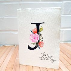 a birthday card with the letter j painted on it and flowers in the middle, sitting on a wooden table