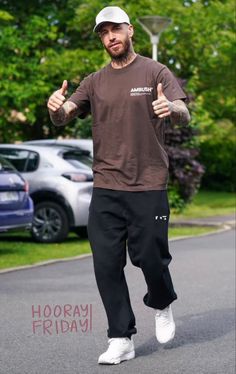 a man in brown shirt and black pants walking down street with his thumb up to the camera