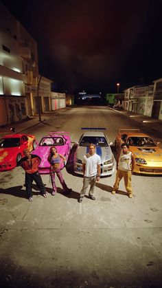 three men and two women standing next to their cars in the middle of an empty street