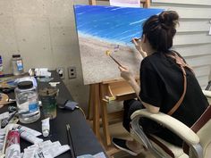 a woman sitting in front of a easel holding a paintbrush and painting an ocean scene