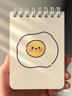 a hand holding a spiral notebook with an egg drawn on it