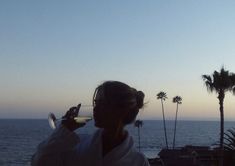 a man drinking from a wine glass next to the ocean with palm trees in the background