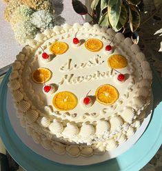 a white cake with oranges and cherries on it sitting on a table next to a potted plant