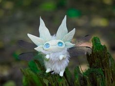 a tiny doll with blue eyes sitting on top of a piece of green wood in the woods