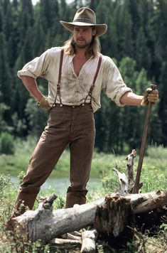 a man standing next to a tree stump with a stick in his hand and wearing a hat