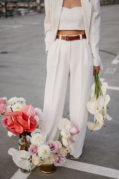 a woman in white is holding flowers and standing next to a vase with pink and white flowers