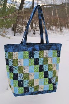 a blue and green tote bag hanging from a tree in the snow with trees behind it