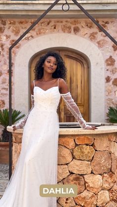 a woman standing in front of a stone wall wearing a white dress and long sleeves