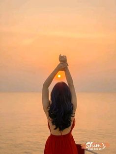 a woman in a red dress is holding her hands up to the sun over the water