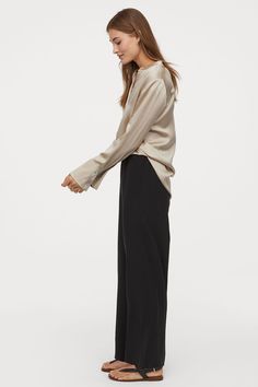 a woman standing in front of a white background wearing a beige sweater and black wide legged pants