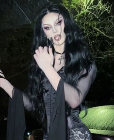 a woman with long black hair and makeup holding something in her hand while wearing gothic clothing