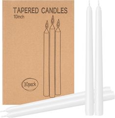 two white candles sitting next to each other in front of a cardboard box with the words tapered candles on it