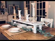 a dining room table with white dishes and candlesticks on it, surrounded by greenery