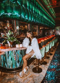 a woman sitting at a bar wearing a white feather jacket and high heeled sandals