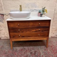 a white sink sitting on top of a wooden dresser next to a brick wall and potted plants