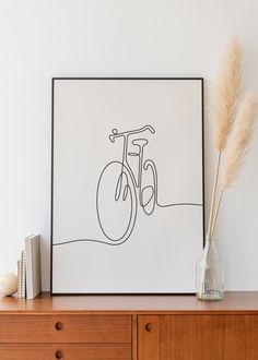 a vase with some dry grass in it next to a framed bicycle print on a dresser