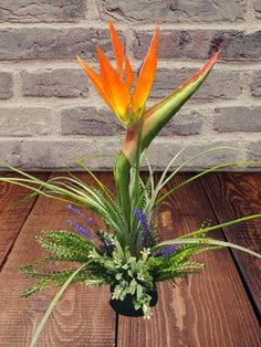 an orange and green flower arrangement in a black vase on a wooden table next to a brick wall