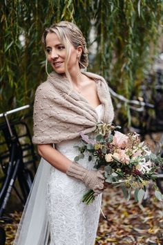 a woman in a white dress holding a bouquet and wearing a knit shawl over her shoulders