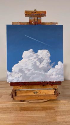 an easel with a painting on it that has clouds and a plane in the sky