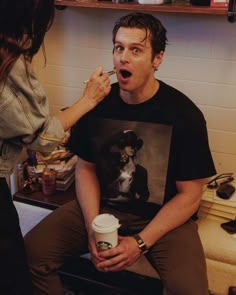 a man sitting on a counter brushing his teeth while holding a coffee cup in front of him