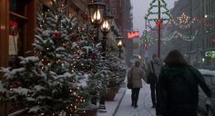 two people walking down a snowy street with christmas lights on the trees and buildings in the background