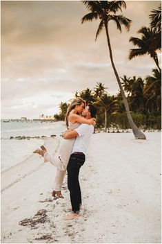 a man and woman hugging on the beach with palm trees in the background at sunset