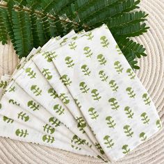 four napkins with green designs on them sitting next to a fern leaf and wicker basket