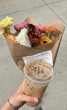 a person holding up a cup of coffee with flowers in the background and a brown paper bag