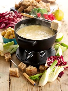 a fonding pot filled with soup surrounded by vegetables and crackers on a wooden table