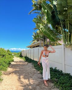 a woman walking down a dirt road next to a white fence and palm trees in the background