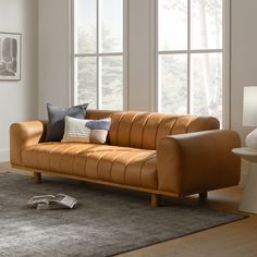 a tan leather couch sitting on top of a wooden floor next to two large windows