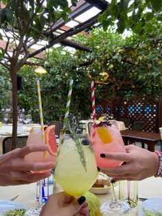 three people are holding up glasses with drinks in them on a table under an umbrella