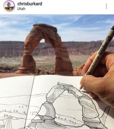 a hand holding a pencil over an open book in front of a desert landscape with arches and rocks